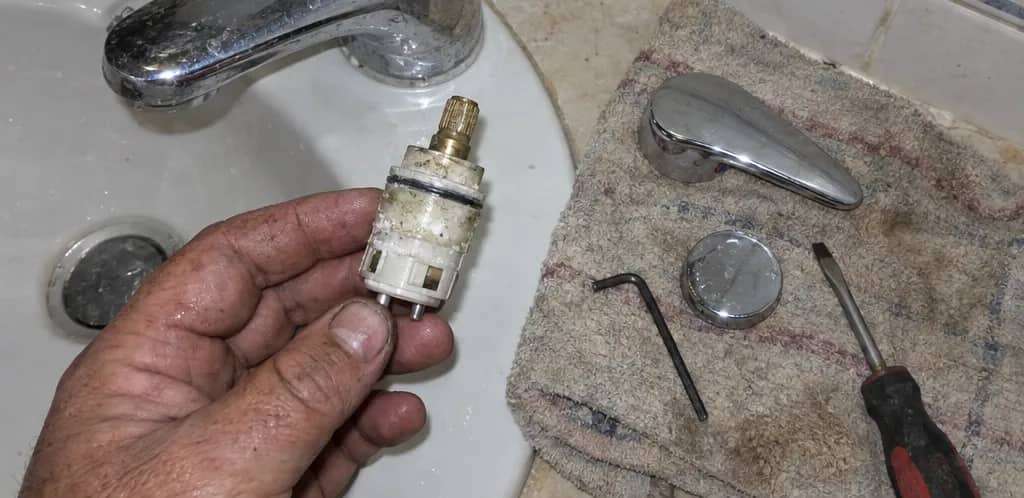 Mano sujetando un cartucho cerámico usado, con restos de suciedad, junto a la maneta, el embellecedor, una llave Allen y un destornillador sobre una toalla