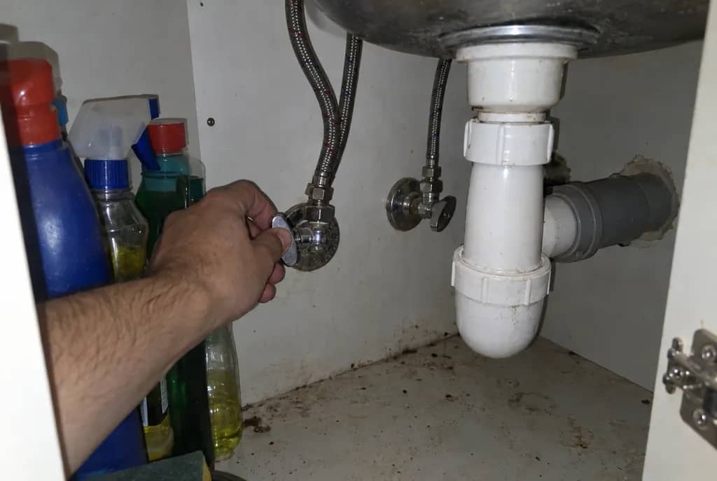 Mano cerrando una llave de escuadra cromada bajo un fregadero, con los latiguillos trenzados subiendo al grifo y el sifón de PVC al lado
